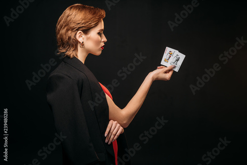 side view of attractive girl in red dress and black jacket holding poker cards isolated on black