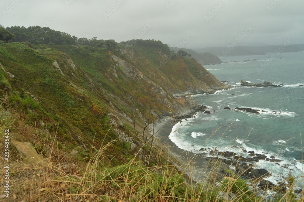 Green Cliffs With Sea Waves Breaking Against Their Rocks At Las Llanas Beach On A Rainy Day. July 29, 2015. Landscapes, Nature, Travel. San Esteban De Pravia, Asturias, Spain. Green Cliffs With Sea Waves Breaking Against Their Rocks At Las Llanas Beach On A Rainy Day. July 29, 2015. Landscapes, Nature, Travel. San Esteban De Pravia, Asturias, Spain.