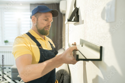 Content concentrated handsome young bearded handyman in cap standing at wall and hanging shelf while working in flat