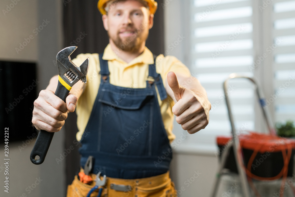 Close-up of positive young handyman showing thumb-up and holding work tool while standing in modern apartment Close-up of positive young handyman showing thumb-up and holding work tool while standing in modern apartment