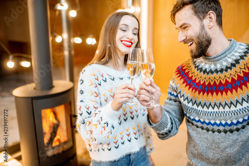 Young couple in sweaters celebrating winter holidays standing with sparkling wine in the modern house with fire place