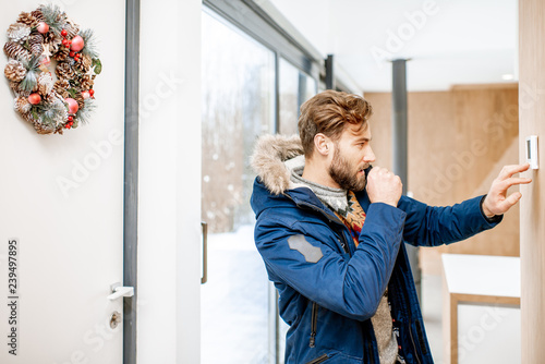 Man in winter clothes feeling cold adjusting room temperature with electronic thermostat at home