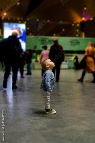 Baby boy standing alone in the airport departure hall in the moving crowd, looking up