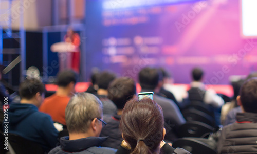 "Audience Watching a Presentation. Business. Female Presenter on Stage Giving Talk to Crowd of People. Woman Speaker at Investor Pitch Conference. Defocused Blurred Presenter During Conference."