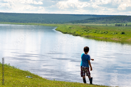 Yakut boy in rubber boots on the precipice above the river at summer sunny day. Back view. Empty space for text