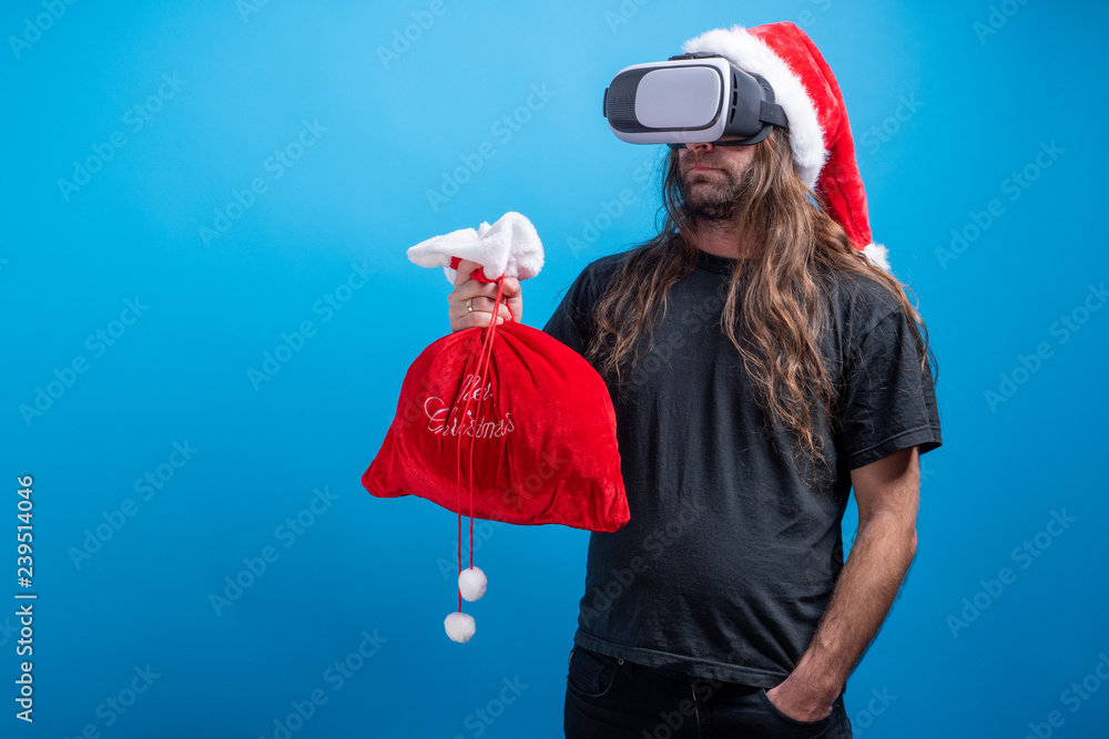 Obraz premium Portrait of male Santa Claus hat and a VR gear, holding bag while having hand in the pocket. Shot in landascape mode. Copy space for advertise