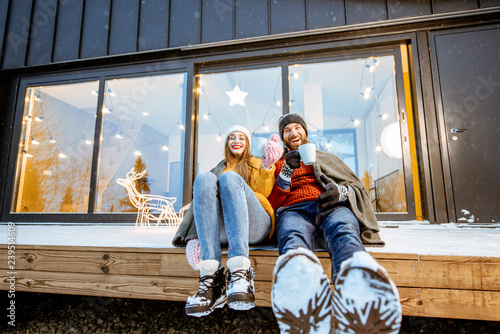 Young couple warming up with plaid and hot drinks sitting on the terrace of the modern house in the mountains durnig the winter holidays