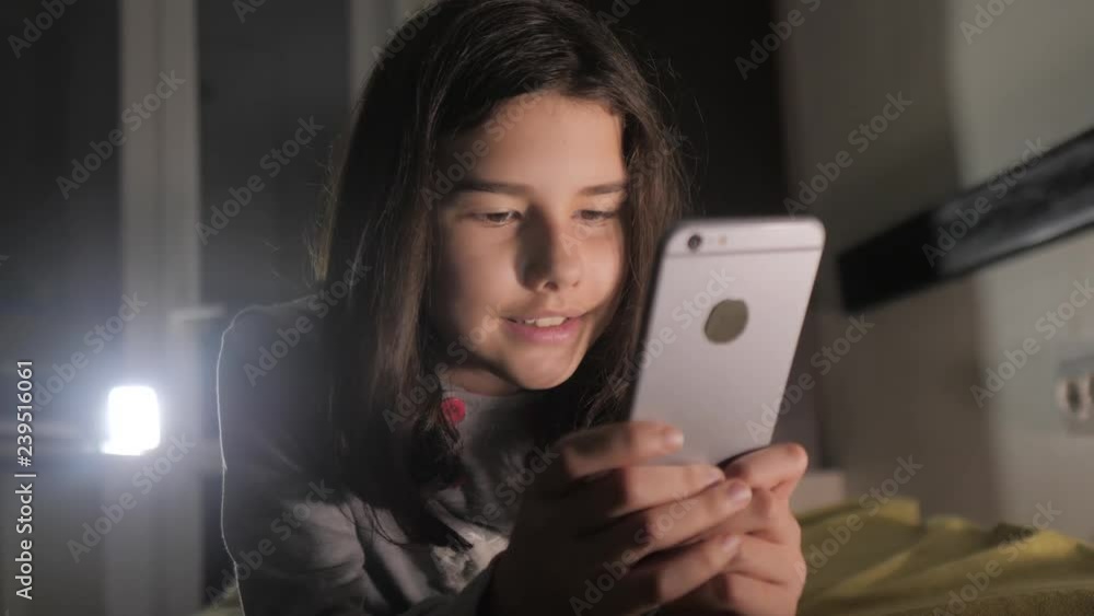 Teenage girl online shopping lifestyle looks on her smartphone in bed during the night. little teen hood girl writes a message chatting in social media messenger night evening indoors smartphone on Teenage girl online shopping lifestyle looks on her smartphone in bed during the night. little teen hood girl writes a message chatting in social media messenger night evening indoors smartphone on