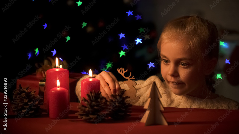Adorable girl playing with wooden Christmas decorations near burning candles Adorable girl playing with wooden Christmas decorations near burning candles