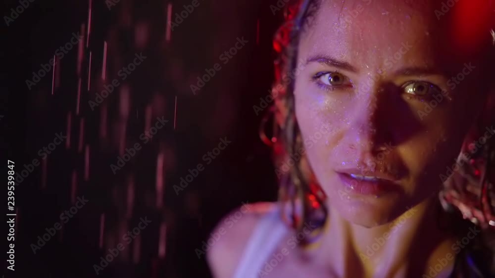 Close-up of a beautiful woman standing under rain indoor of a studio, pink light falling on her face. Close-up of a beautiful woman standing under rain indoor of a studio, pink light falling on her face.