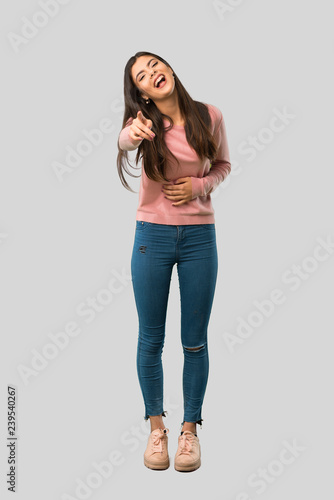 Full body of Teenager girl with pink shirt pointing with finger at someone and laughing a lot on isolated grey background