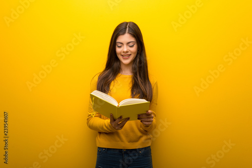 Teenager girl on vibrant yellow background holding a book and enjoying reading