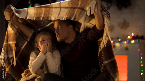 Father telling horror Christmas story for little girl sitting under cozy plaid