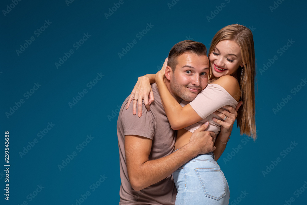 Young couple is hugging on blue background in studio. They wear T-shirts, jeans and smile. Young couple is hugging on blue background in studio. They wear T-shirts, jeans and smile.