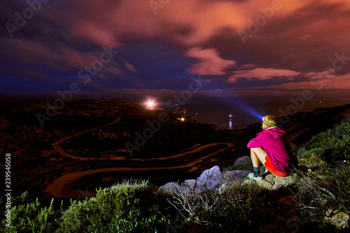 Tourist girl sitting on a rock over the sea and cliffs of Corsica Corse France and watching the night scenery   clouds and the lighthouse in the distance