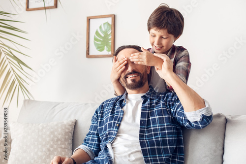 Father and little son at home boy covering eyes of man sitting on sofa making surprise happy