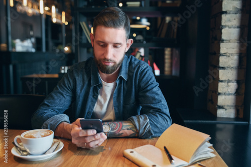Young bearded businessman,dressed in a denim shirt, sitting at table in cafe and use smartphone. Man using gadget.