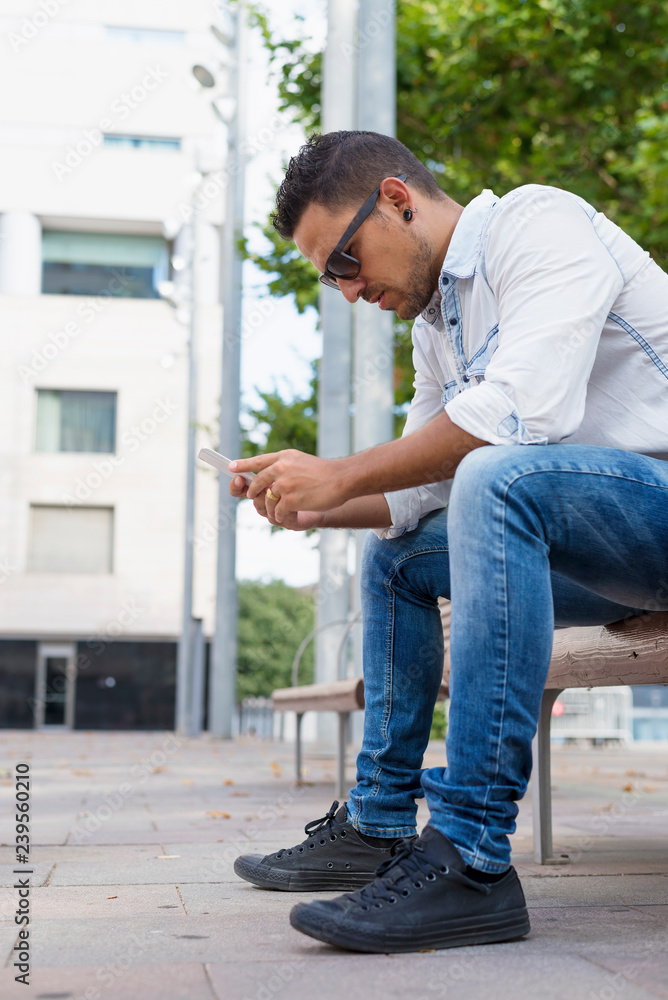 Young man wearing sunglasses using a mobile while sitting on a bench Young man wearing sunglasses using a mobile while sitting on a bench