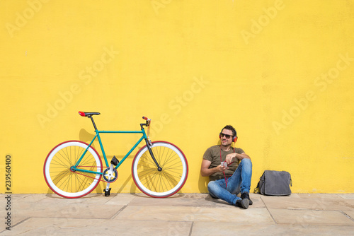 Young man wearing sunglasses relaxing on the floor and listening music. Sitting against a yellow wall with his computer
