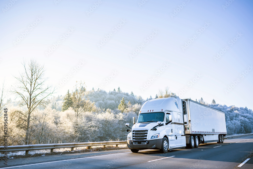 Big rig white technological semi truck with reefer semi trailer going on the winter road with frost trees on the hill