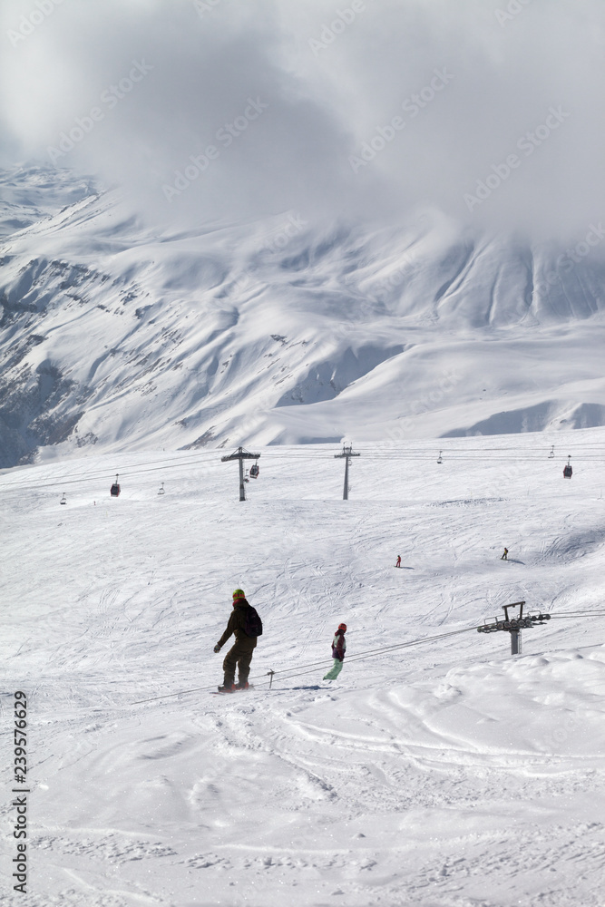 Snowboarders on snowy ski slope and high mountains in gray cloud