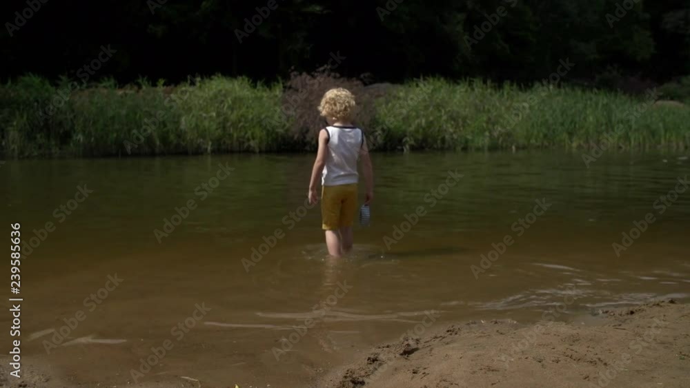5-year-old blond-haired kid goes into river and lowers paper boat into water. 5-year-old blond-haired kid goes into river and lowers paper boat into water.