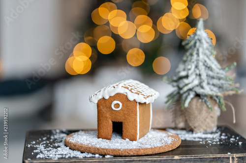 Gingerbread house on wooden table. Defocused garland lights on background. Christmas tree and Holiday mood. Morning in the bright living room.