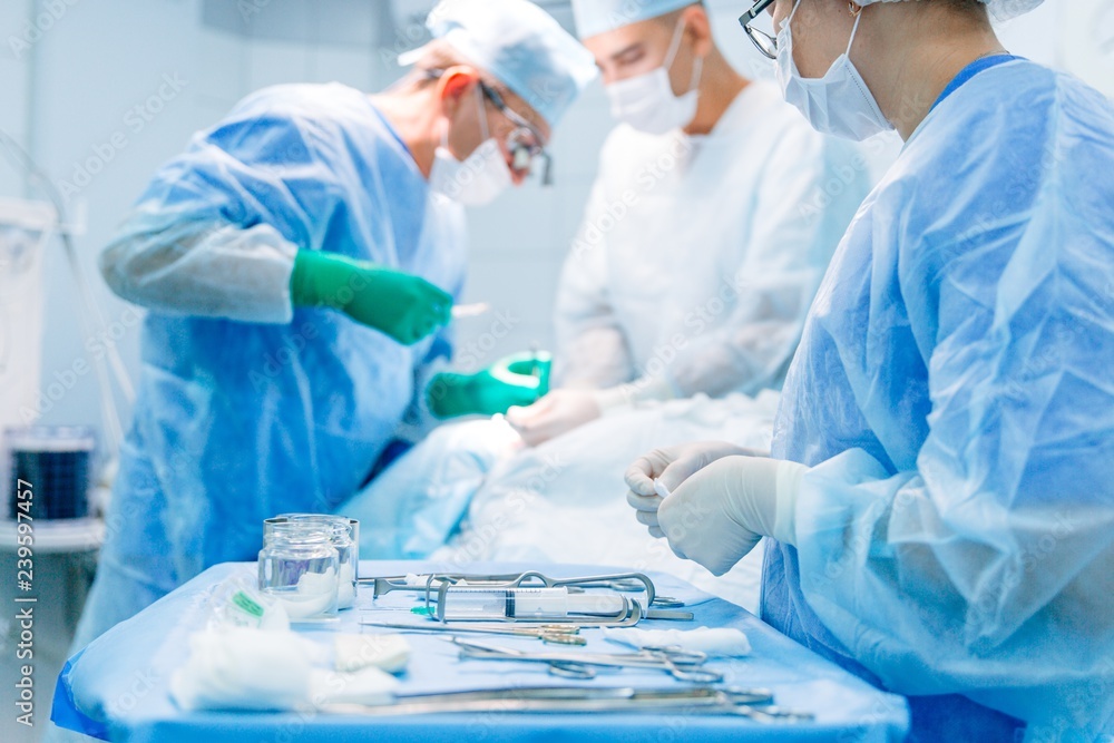 Surgeon working with a scalpel, wearing a blue surgical mask and a surgical cap in intensive care with his team of surgeons Surgeon working with a scalpel, wearing a blue surgical mask and a surgical cap in intensive care with his team of surgeons