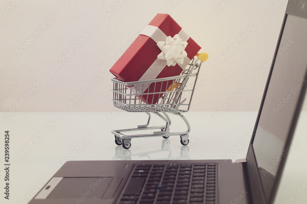 Small trolley with a red gift and a black computer. Buy products on an ecommerce in internet. Last minute christmas shopping online concept.