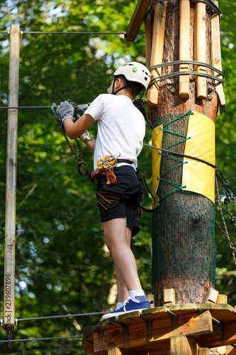 Child in forest adventure park. Kid in white helmet and white t shirt climbs on high rope trail. Agility skills and climbing outdoor amusement center for children. young boy plays outdoors