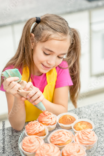 Smiling little girl with confectionery bag squeezing  cream on cupcakes at kitchen