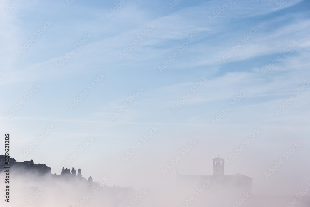 A view of a silhouette of St.Francis church in Assisi in the middle of mist beneath a blue sky with clouds