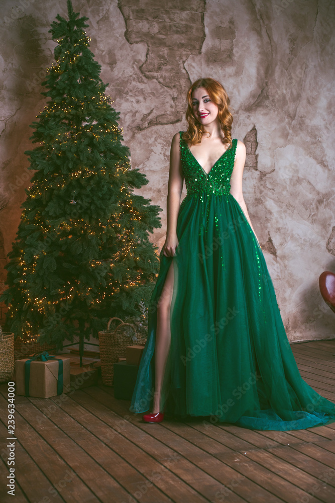 Fototapeta premium Winter holidays, celebration and people concept - young sexy woman in elegant green evening dress over christmas interior background