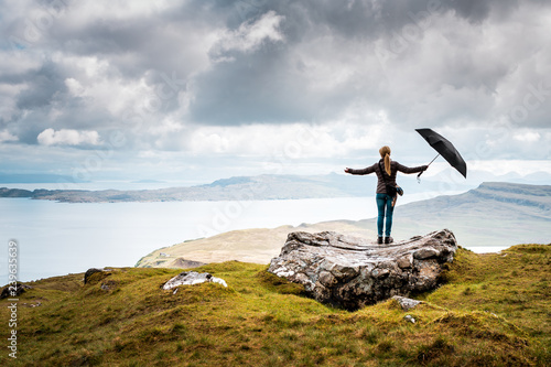 Woman stands on big rock with arms wide open and an umbrella in her hand, rainy weather in the scottish highlands, light and hope, after the rain