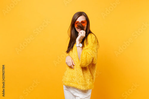 Smiling funny young woman in fur sweater and heart orange glasses holding, covering face with hair isolated on bright yellow background. People sincere emotions, lifestyle concept. Advertising area.