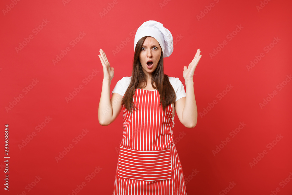 Housewife female chef cook or baker in striped apron, white t-shirt, toque chefs hat isolated on red wall background. Beautiful shocked housekeeper woman spreading hands. Mock up copy space concept. Housewife female chef cook or baker in striped apron, white t-shirt, toque chefs hat isolated on red wall background. Beautiful shocked housekeeper woman spreading hands. Mock up copy space concept.