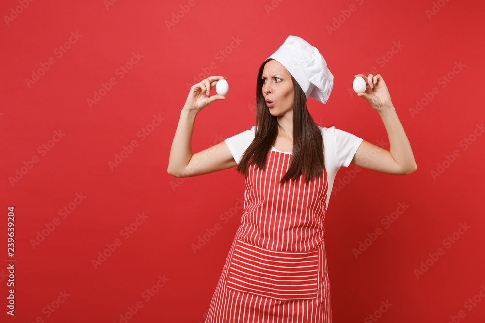 Fototapeta premium Housewife female chef cook or baker in striped apron, white t-shirt, toque chefs hat isolated on red wall background. Fun housekeeper woman hold in hands two chicken eggs. Mock up copy space concept.