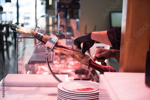 Preparation of a jamon meat from the leg is seen on the picture. The hands of the worker is seen wearing black colored gloves. White dishes are seen too.