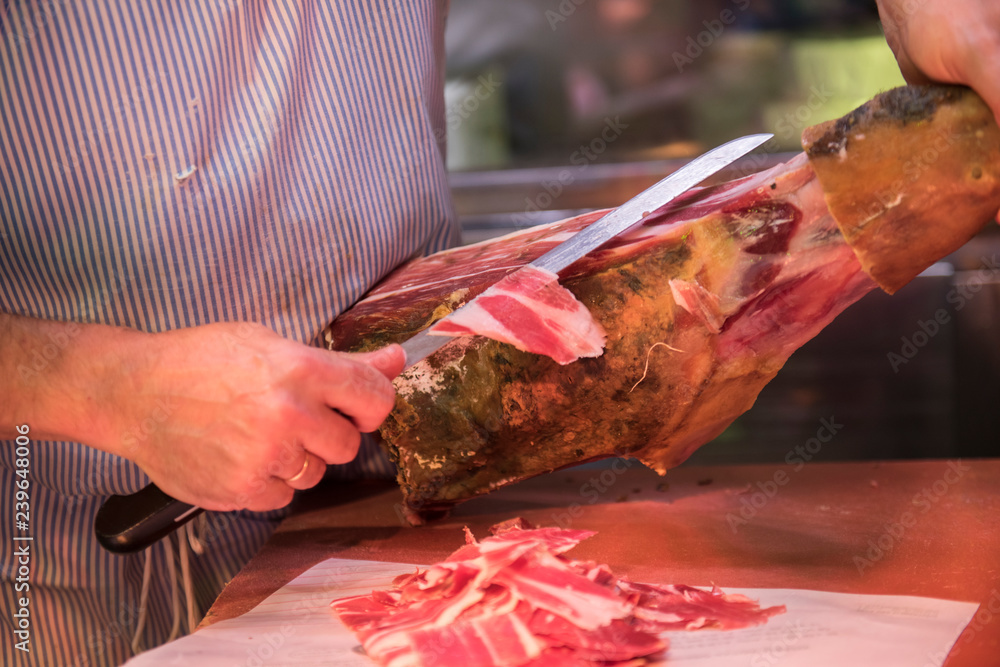 A butcher is seen cutting jamon meat with a butcher knife. The jamon looks fresh and is ready to be sold after being cut. He is seen wearing a stripe Apron.