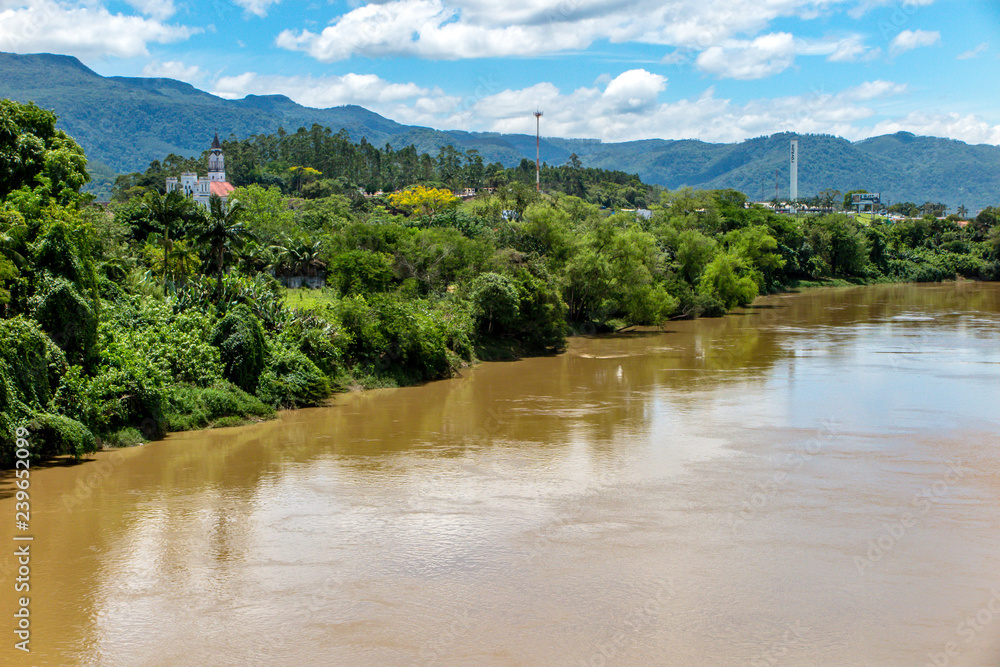 Muddy river with lots of vegetation next to the city of Ascura, mountains with forest in the background, blue sky with clouds, European Valley, Santa Catarina