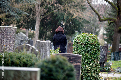 Trauernde Frau vor Grab auf Friedhof mit gefalteten Händen als Rückenansicht.
