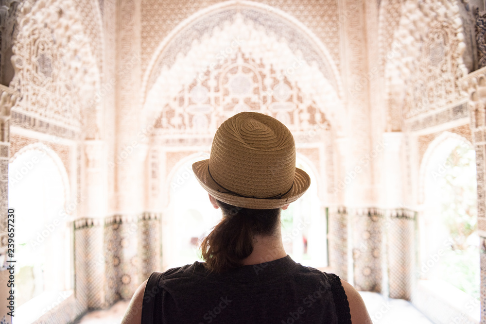 A female tourist is seen standing at the top of Alhambra Palace and looking outside through the balcony. The lady is seen wearing a white hat.
