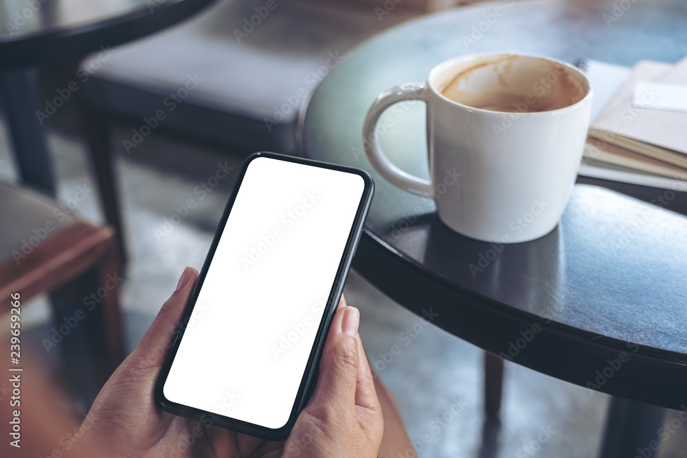 Mockup image of woman's hands holding and using a black mobile phone with blank screen with coffee cup on table