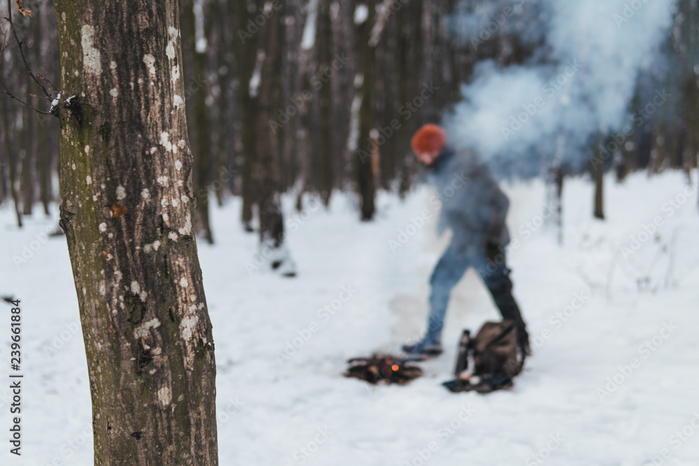 Traveling man makes a fire. Winter time. Concept adventure active vacations outdoor. Extreme camping. vintage backpack and thermos with tea, snowy forest