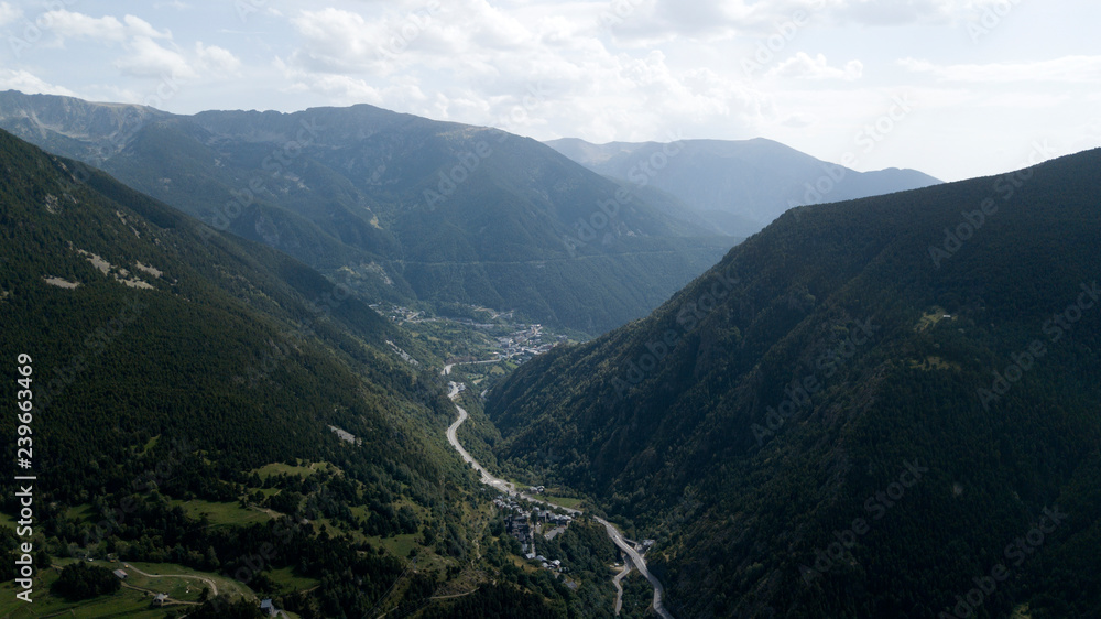 Aerial image of the green valley in the Pyrenees mountains in the summer. There are green mountains with trees down below at Andorra La Vella, Andorra. There are city with brown buildings down below.