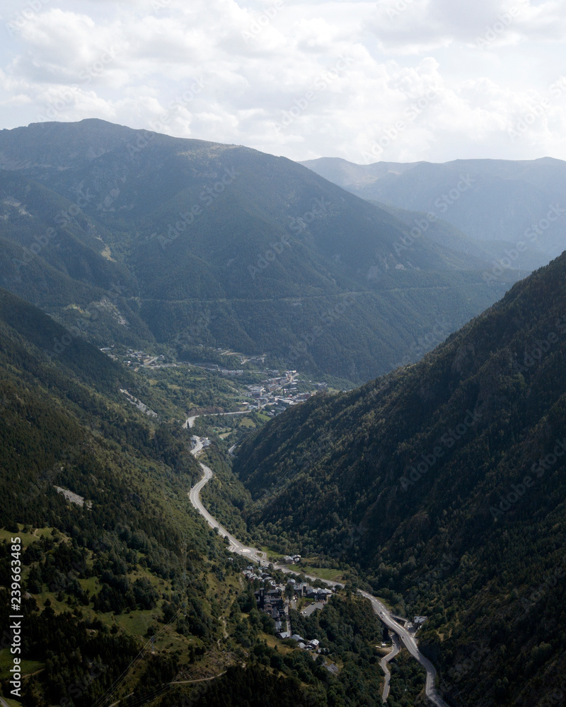 Aerial photo of the green valley in Pyrenean mountains in the summer. There are green mountains with trees and rocks down below at Andorra La Vella, Andorra. There are city with brown buildings and Aerial photo of the green valley in Pyrenean mountains in the summer. There are green mountains with trees and rocks down below at Andorra La Vella, Andorra. There are city with brown buildings and