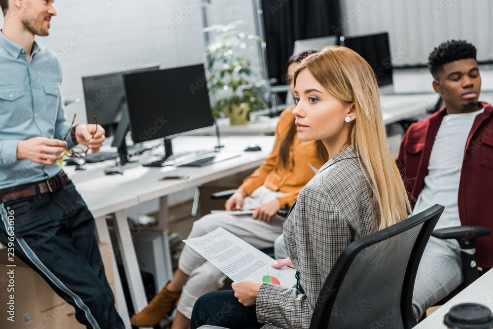 selective focus of young businesswoman with papers near multiracial colleagues at workplace in office selective focus of young businesswoman with papers near multiracial colleagues at workplace in office