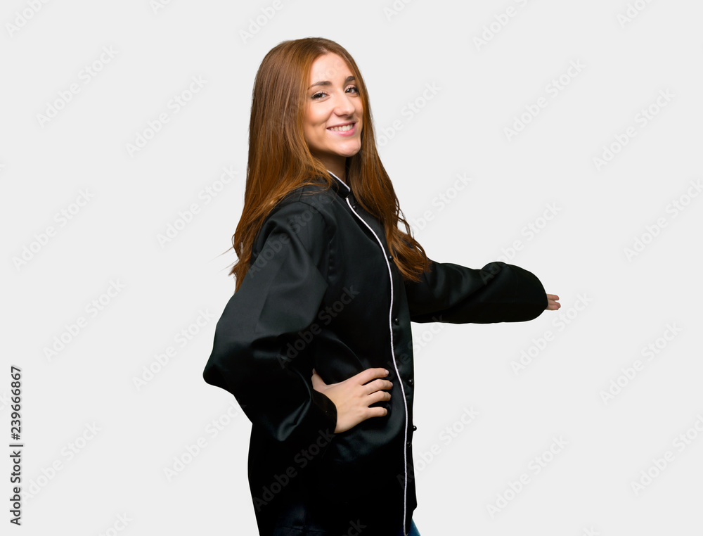 Young redhead chef woman pointing back and presenting a product on isolated grey background Young redhead chef woman pointing back and presenting a product on isolated grey background
