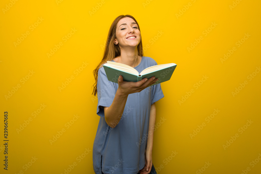 Young redhead girl over yellow wall background holding a book and giving it to someone Young redhead girl over yellow wall background holding a book and giving it to someone