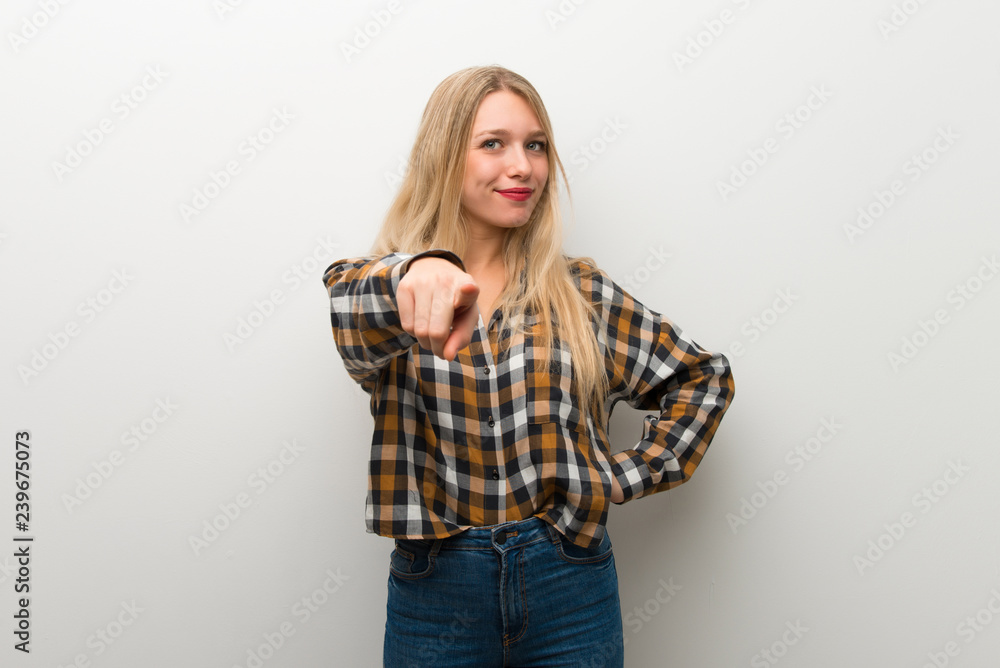 Blonde young girl over white wall points finger at you with a confident expression Blonde young girl over white wall points finger at you with a confident expression